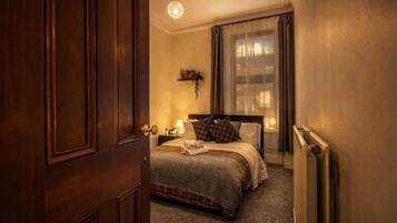 Una habitación de hotel con puerta de madera, cama con cubrecama a cuadros, mesita de noche con lámpara y ventana con cortinas traslúcidas.