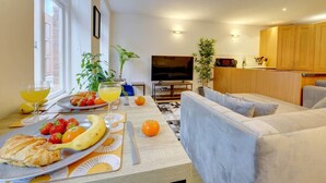 Un área de comedor con una mesa lista para el desayuno, que incluye frutas y pasteles, y un sofá al fondo.
