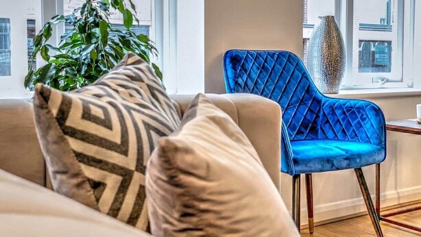Una sala de estar moderna con un sofá color beige, un sillón de terciopelo azul y un cojín estampado.