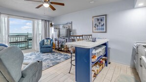 Ein geräumiges Wohnzimmer mit großem Fenster zum Meer, einem Deckenventilator, einer Essecke mit Kücheninsel und einem blauen Sofa.