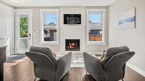 A living room with a fireplace, two grey armchairs, and a view of the outside through large windows.