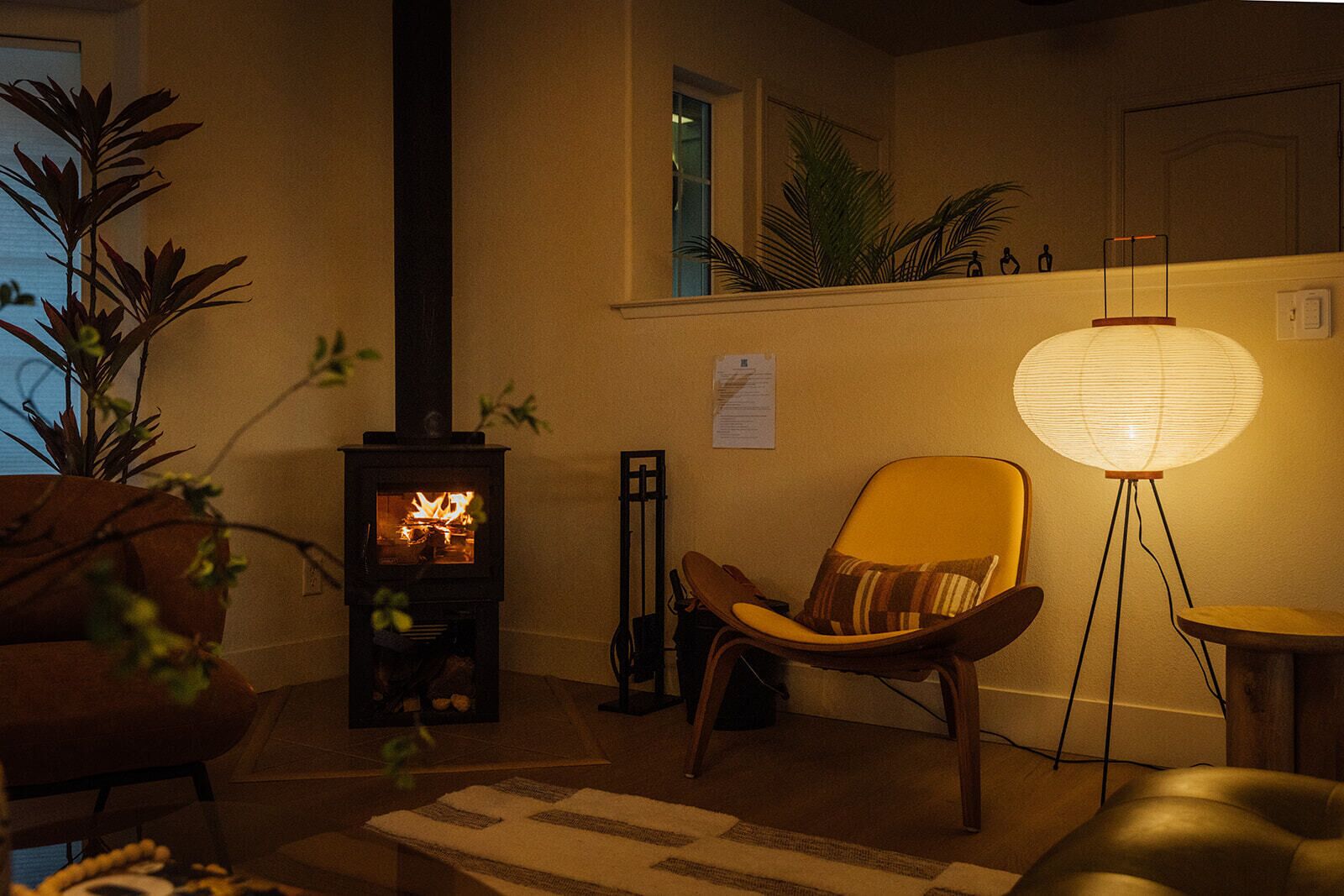 A cozy living room with a wood-burning stove, a comfortable chair, a floor lamp, and a potted plant.