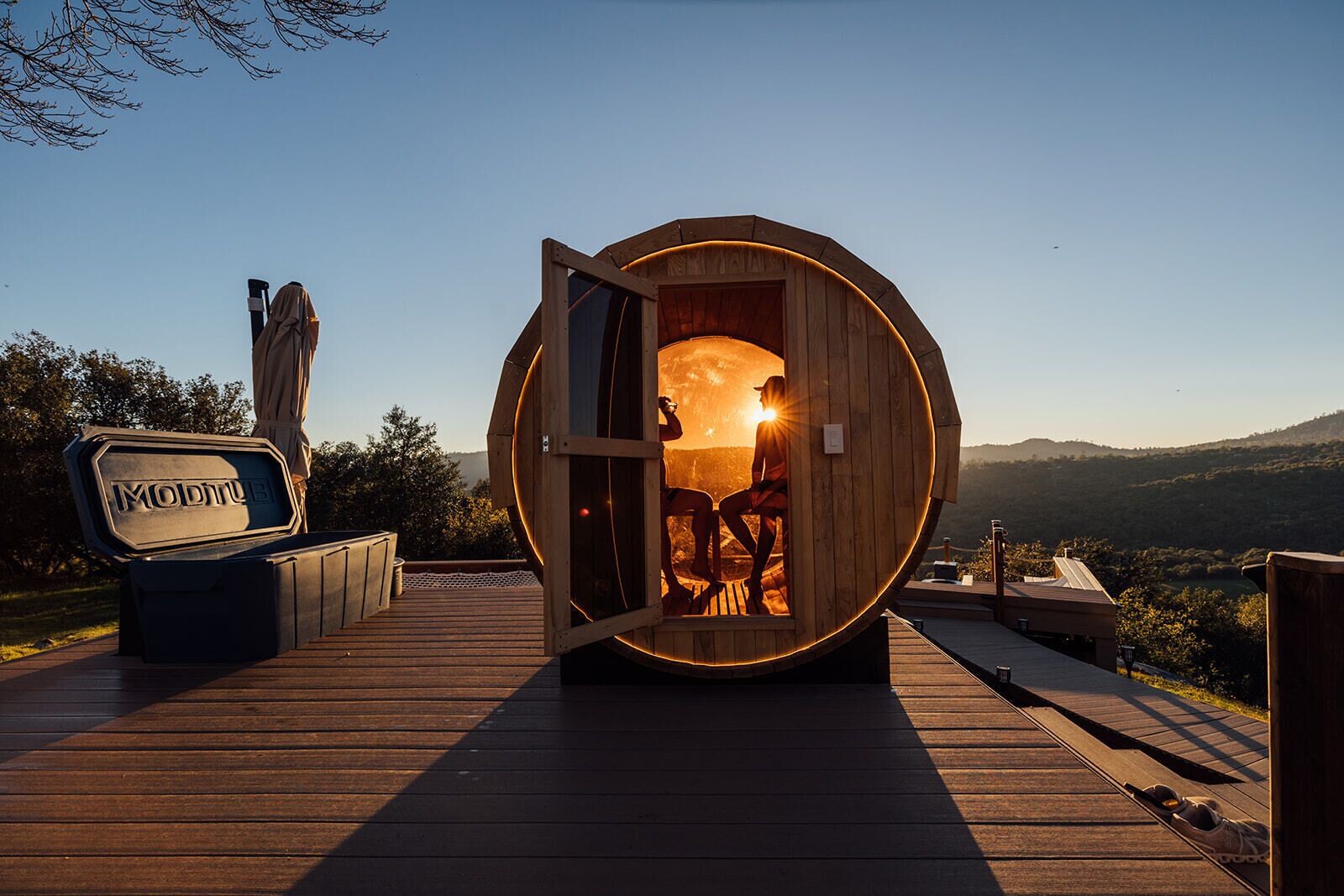 Eine Holzsauna mit geöffneter Tür, in der zwei Personen zu sehen sind, steht auf einer Holzterrasse mit herrlicher Aussicht.