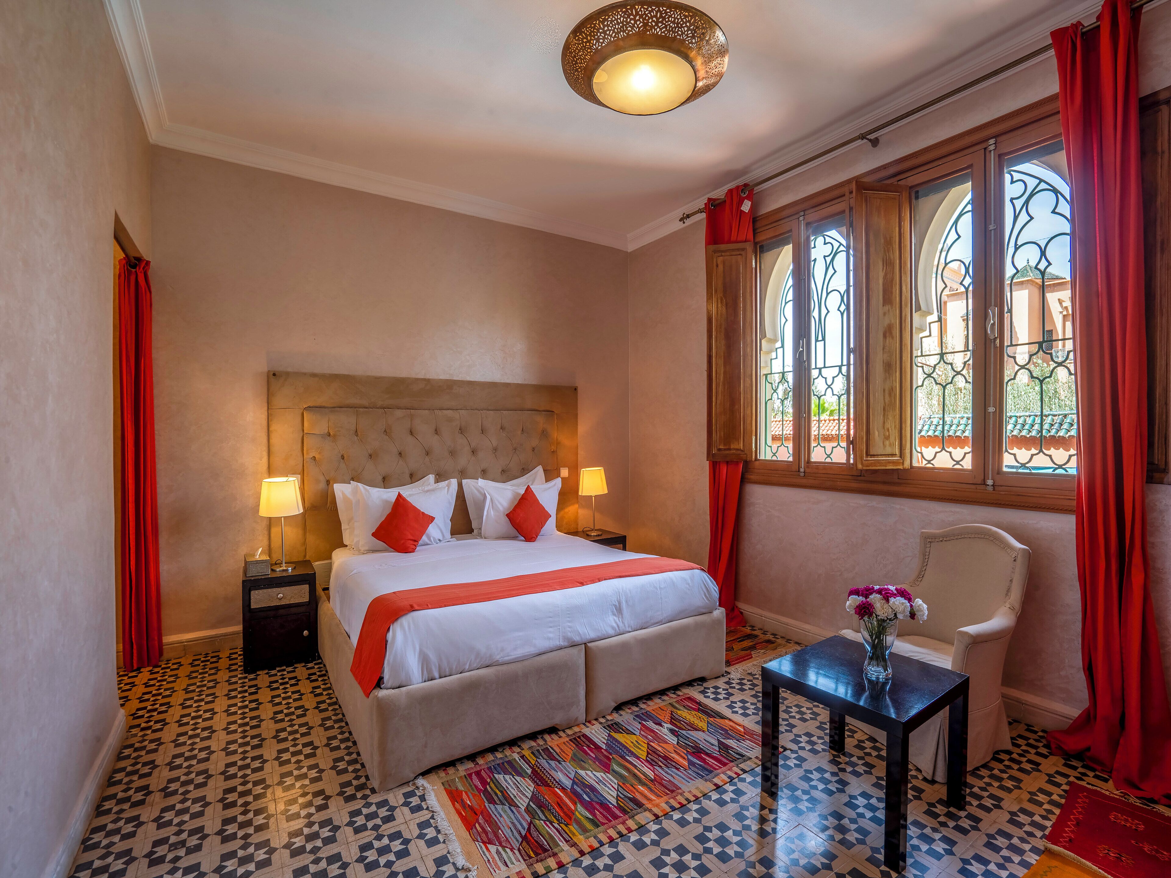 A hotel room with a large bed, two bedside tables with lamps, a chair, a small table with a vase, patterned floor tiles, and a window with red curtains.