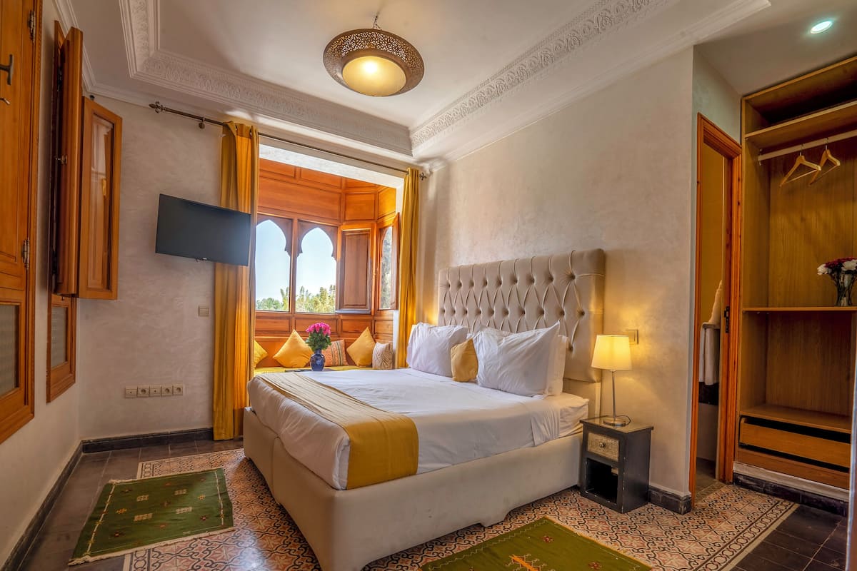 A hotel room with a large bed, a television, a window with curtains, a wardrobe, and a patterned floor.