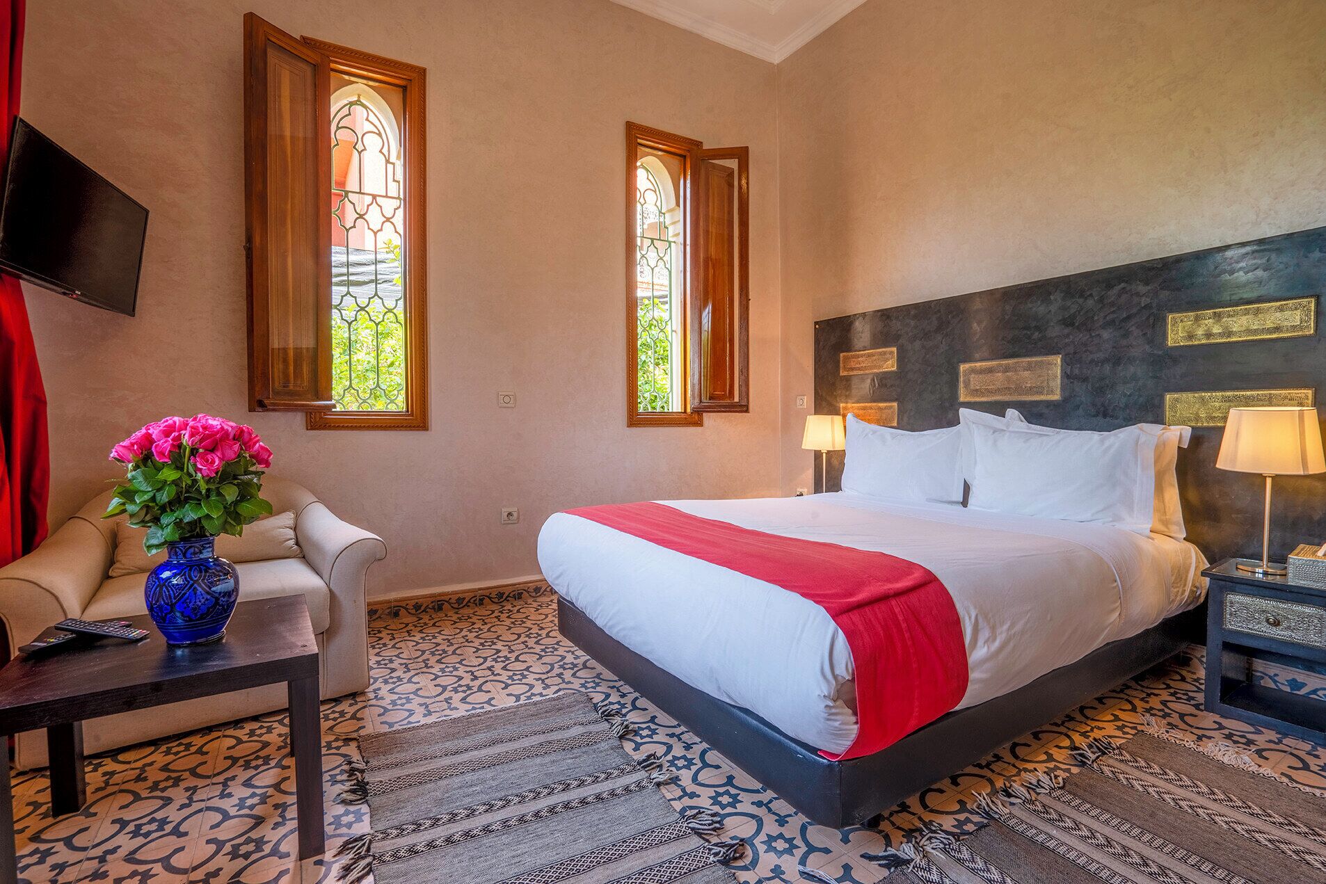 Une chambre d’hôtel avec un grand lit, deux fenêtres ornées de vitraux, un téléviseur fixé au mur, un vase de fleurs posé sur une table d’appoint et une moquette à motifs.