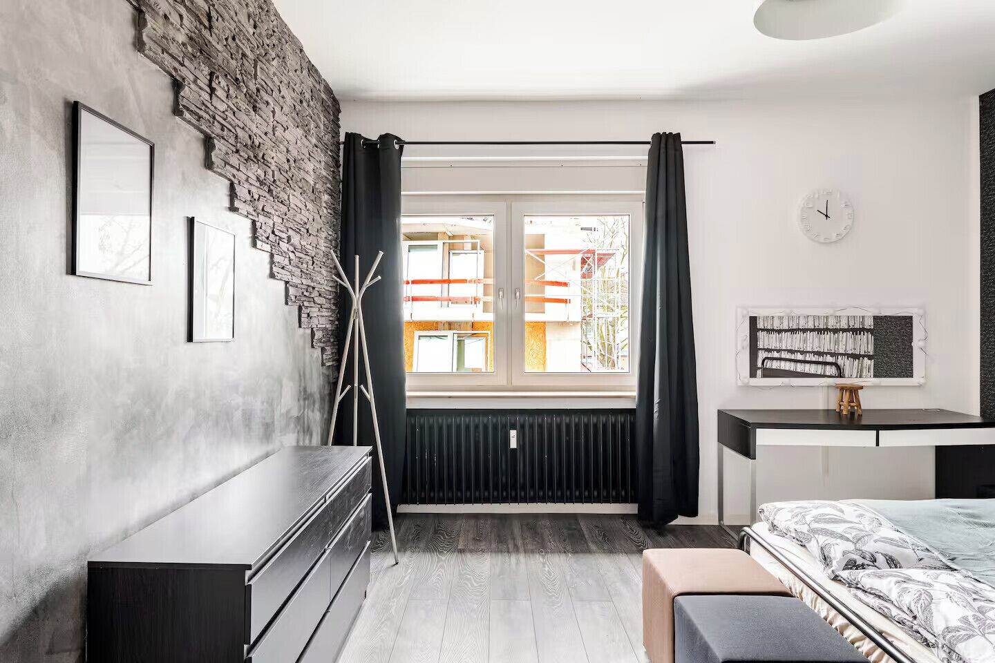 Een moderne slaapkamer met een accentmuur van baksteen, een groot raam met gordijnen, een radiator, een bed met een beddeken met een patroon, een nachtkastje en een klok aan de muur.