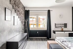 Een moderne slaapkamer met een accentmuur van baksteen, een groot raam met gordijnen, een radiator, een bed met een beddeken met een patroon, een nachtkastje en een klok aan de muur.
