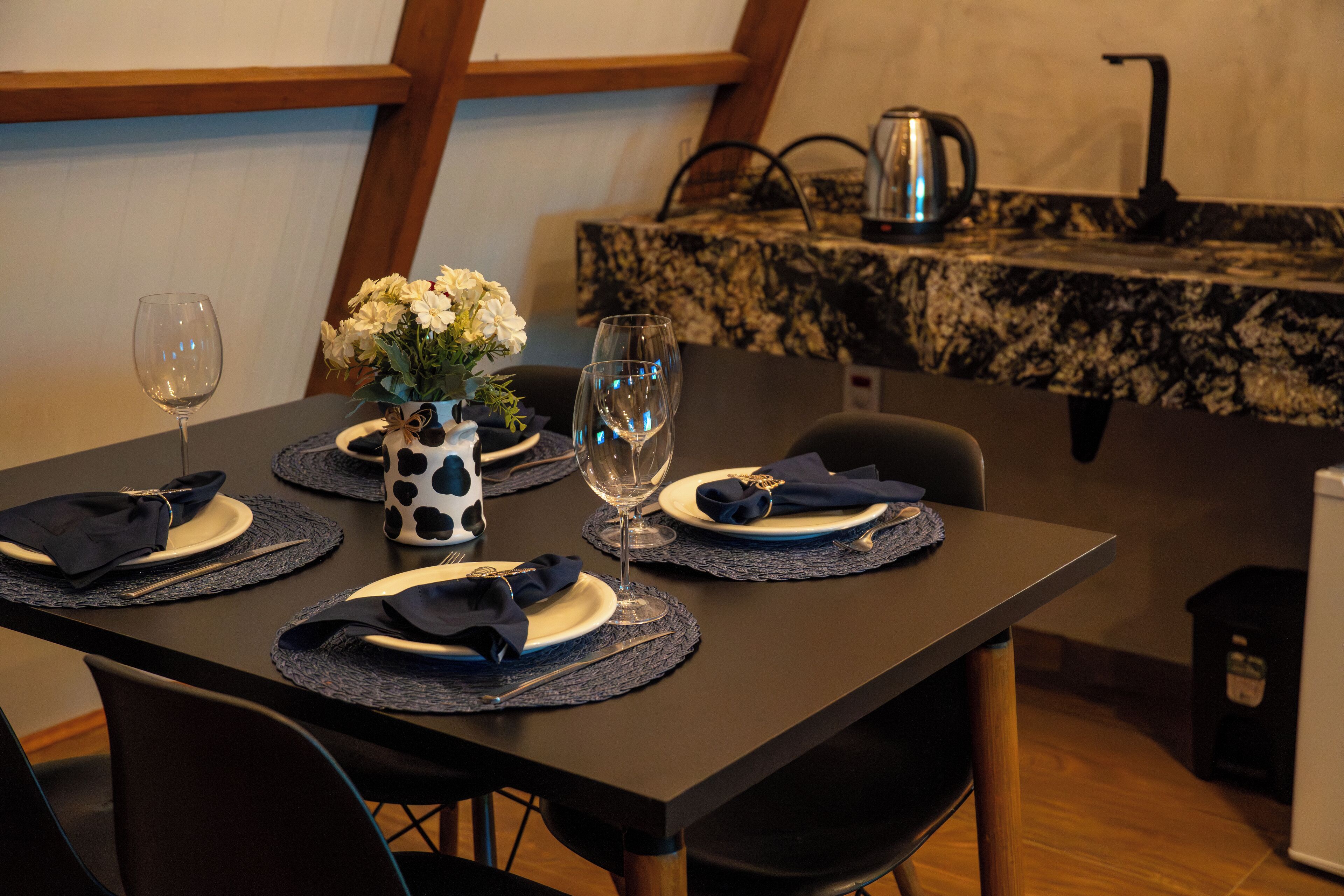Uma mesa de jantar posta com pratos, guardanapos e copos de vinho, com um vaso de flores e uma cafeteira ao fundo.