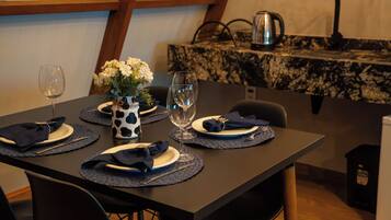 Uma mesa de jantar posta com pratos, guardanapos e copos de vinho, com um vaso de flores e uma cafeteira ao fundo.