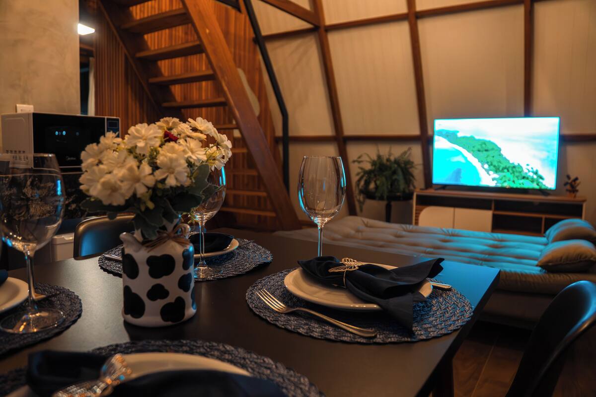 A dining area with a table set for two, a vase of flowers, and a television displaying a scenic view.
