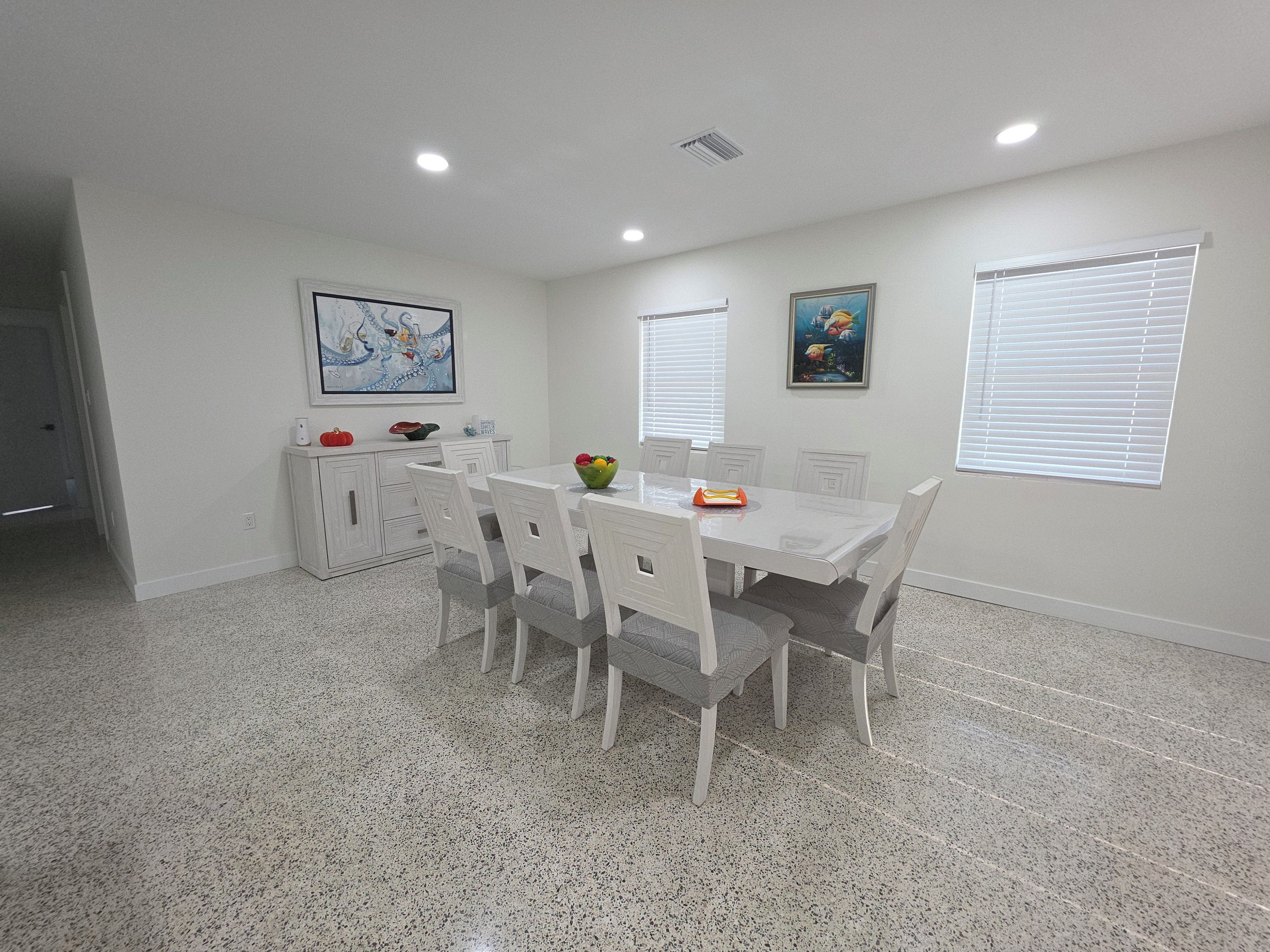 A dining area with a white table and chairs, a fruit bowl, and artwork on the walls.