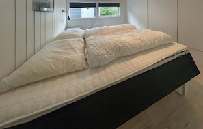 Una cama individual con colchón acolchado y cabecera negra en una habitación con paredes blancas y ventana con persianas.