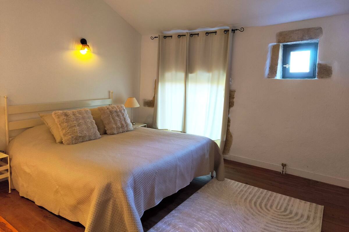 A bedroom with a bed, a nightstand, a lamp, a window with curtains, and a small skylight.