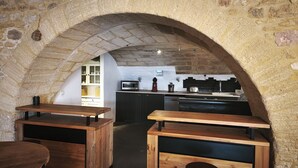 Une cuisine rustique aux murs de pierre, aux tables en bois et dotée d’un îlot central équipé d’appareils électroménagers modernes.
