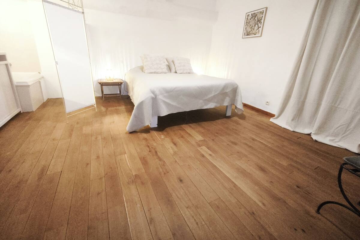 A bedroom with a wooden floor, a bed with a white bedspread, a nightstand, and a wall-mounted picture.