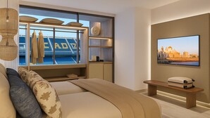 A modern hotel room with a large bed, a television, a wooden bench, and a view of a cityscape through a window. - Gran Hotel Luna de Cádiz *NEW* (Cádiz)