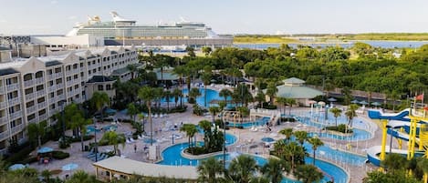 A resort with multiple swimming pools, a large cruise ship in the background, and lush greenery.