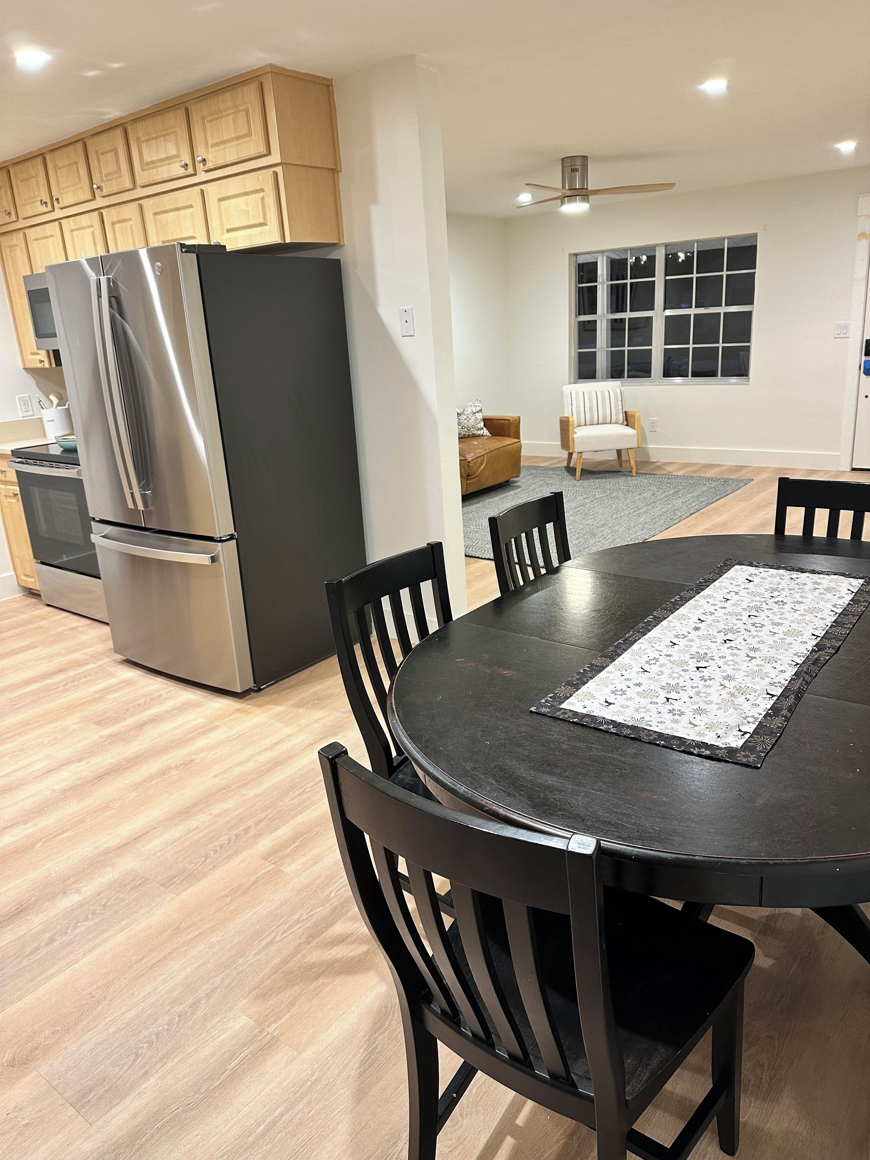 A modern kitchen with stainless steel appliances, a dining area with a round table and chairs, and a living space with a sofa and a window.