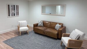 A living room with a brown leather sofa, two white chairs, a grey patterned rug, a ceiling fan, and a large mirror on the wall. - Cozy 3/2 in downtown quaint Lake Mary, FL with AC and fenced in yard (Lake Mary)