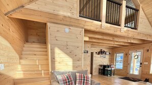A cozy cabin interior with a staircase leading to a loft area, a grey sofa with plaid cushions, and a wooden floor. - Four seasons, waterfront, private beach! Elegant cozy cabin lakeside retreat! (Smithfield)