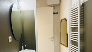 Une salle de bain avec un meuble-lavabo en bois, un lavabo blanc, un miroir, un porte-serviettes et une porte donnant sur l’espace de la douche.