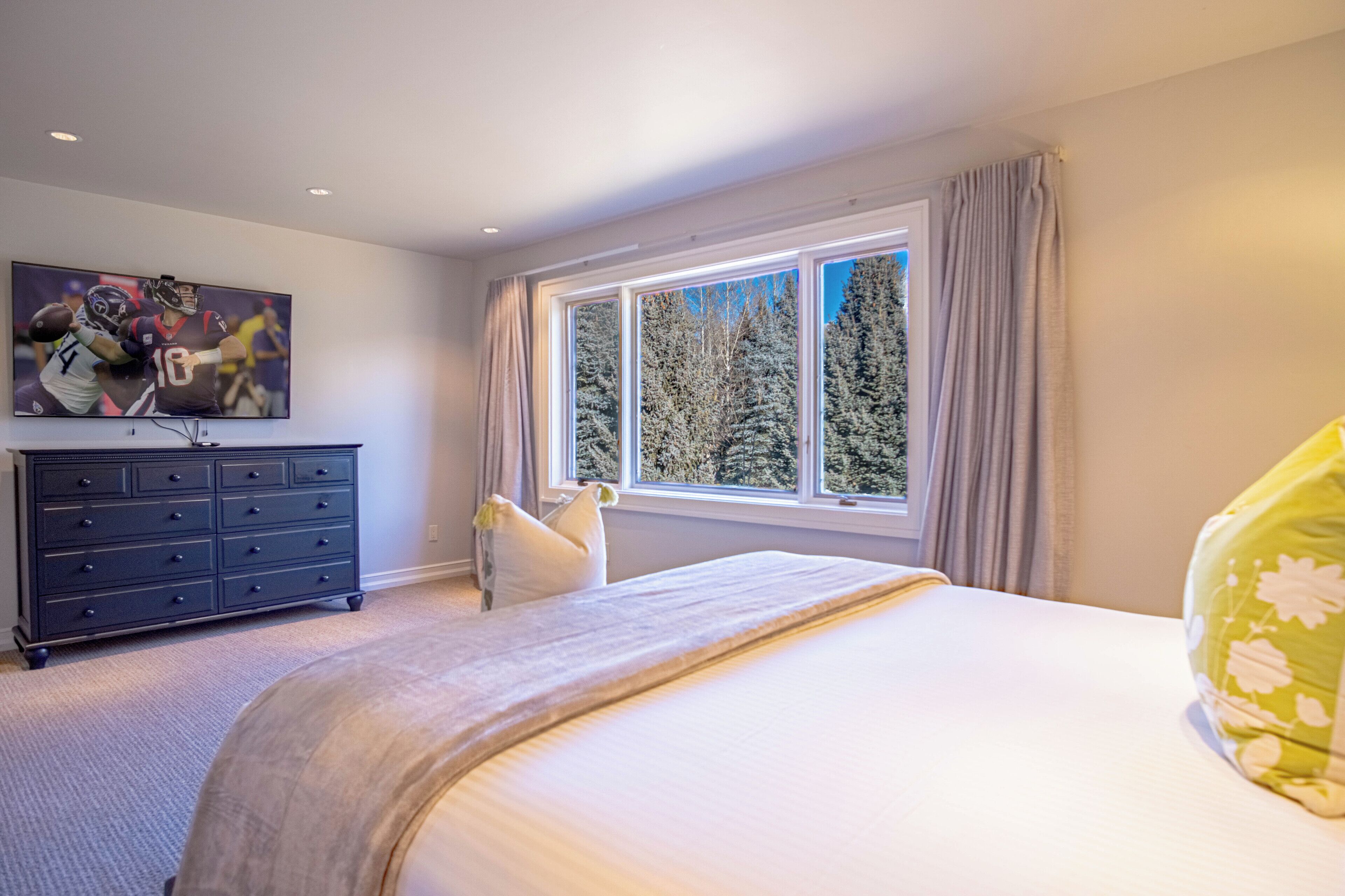 Une chambre avec un grand lit, une commode, un tableau au mur et une fenêtre donnant sur des arbres.