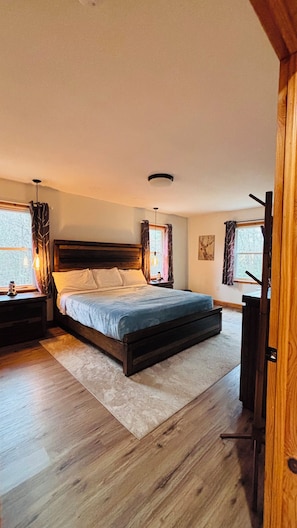Ein Schlafzimmer mit einem großen Bett, einem Holzkopfende, zwei Nachttischen, einem Schreibtischchen, einer Deckenleuchte und einem Fenster mit Vorhängen.