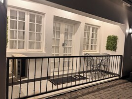 A building with multiple windows, a black metal fence, a small outdoor seating area with chairs, and a green plant decoration.
