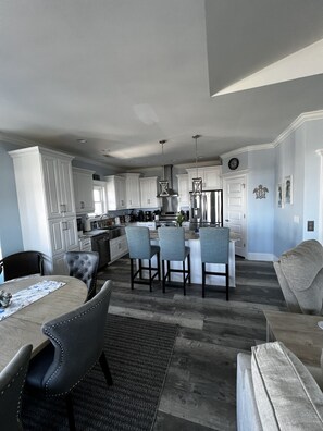 A modern kitchen with white cabinets, a central island with bar stools, and a dining area with a table and chairs. - Topsail Beach Sunsets, Water views, Pool/Hot tub (N. Topsail Beach)