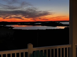 A sunset view from a balcony with a railing, overlooking a body of water and a distant shoreline. - Topsail Beach Sunsets, Water views, Pool/Hot tub (N. Topsail Beach)