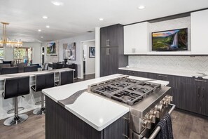 Une cuisine moderne dotée d’un îlot central, de tabourets de bar, de meubles de rangement aux couleurs sombres et d’un grand téléviseur à écran plat placé au-dessus de l’îlot.