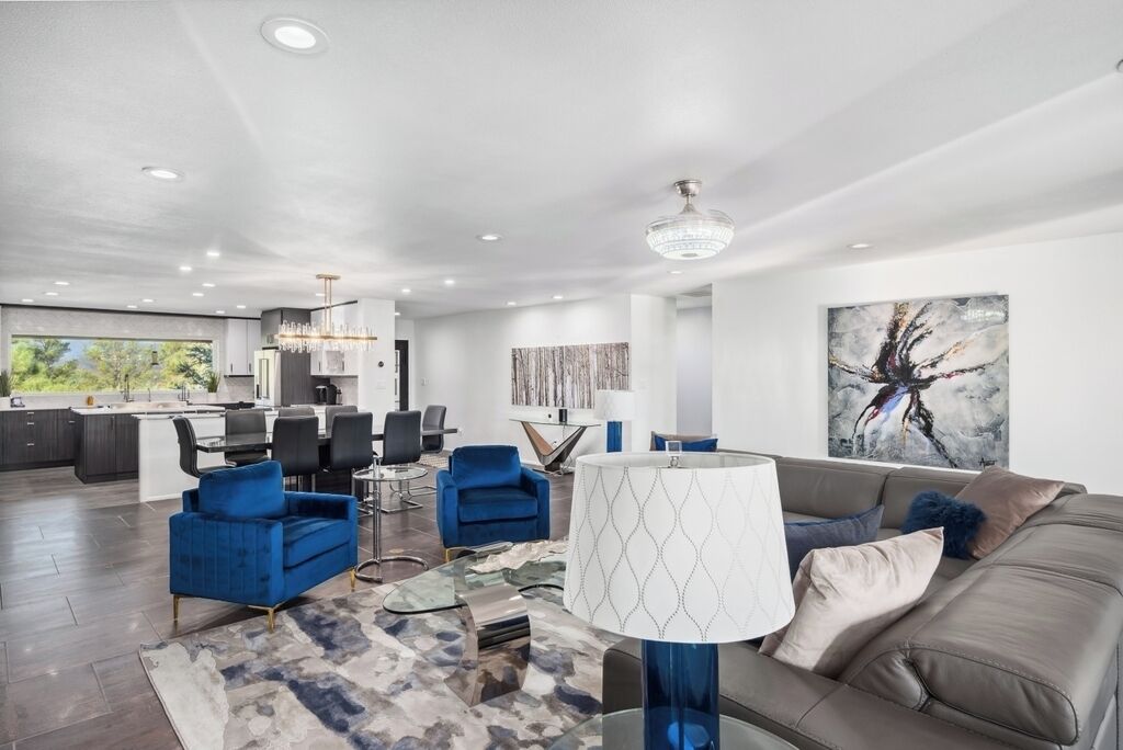 A modern living room with a sofa, blue armchairs, a glass coffee table, and a chandelier. There is a dining area with a table and chairs, and a kitchen in the background.