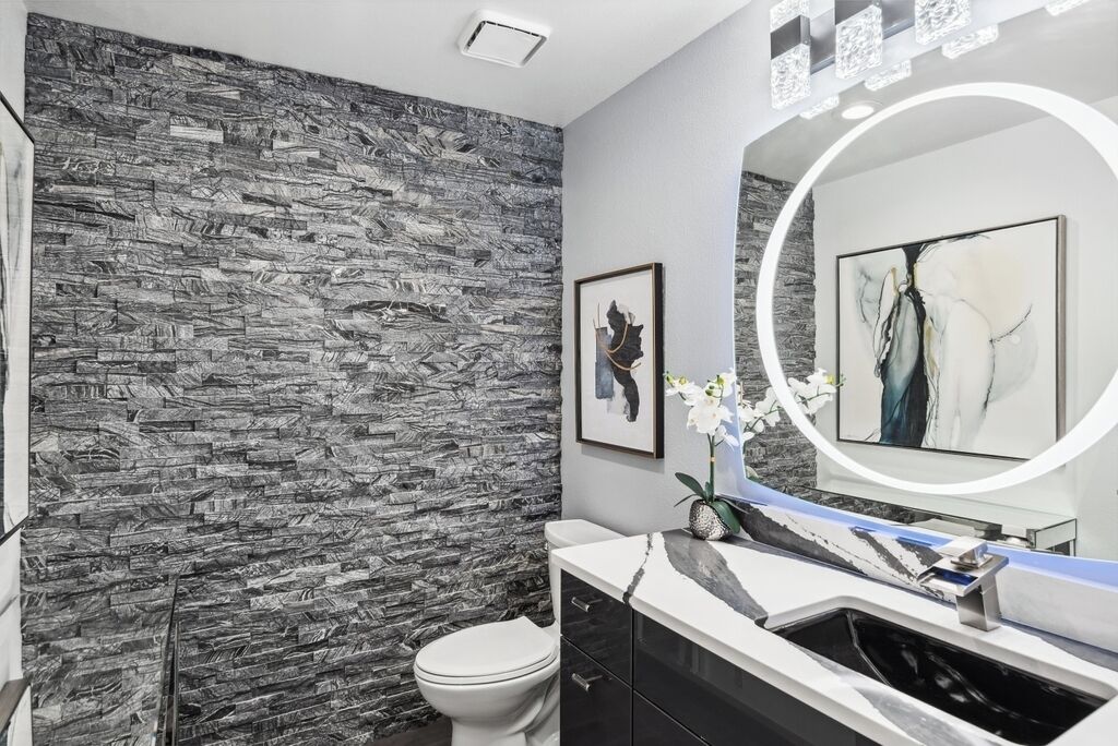 Une salle de bain avec un mur à l’aspect pierre, des toilettes blanches, un lavabo noir doté d’un plan de travail en marbre et un grand miroir avec un éclairage circulaire.