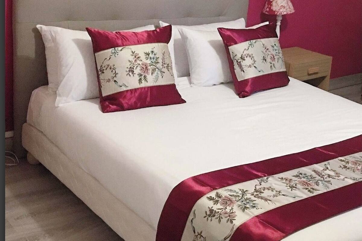 A neatly made bed with white and red bedding, a floral-patterned pillow, and a mirror with a gold frame on a pink wall.