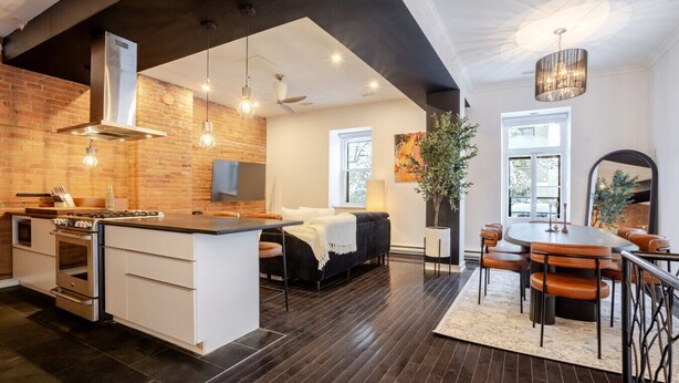 A modern kitchen with a central island, a dining area with a round table, and a living space with a sofa and a large plant. - Elegant Downtown Stay | Near Bell Centre MTL (Montreal)