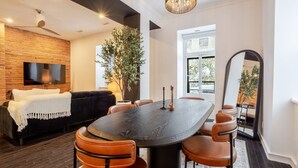 A modern living room with a dark wood dining table, leather chairs, a black sofa, a TV mounted on a brick wall, and a large mirror reflecting the room. - Elegant Downtown Stay | Near Bell Centre MTL (Montreal)