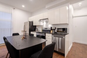A modern kitchen with white cabinets, a black countertop, stainless steel appliances, and a dining area with a dark wooden table and chairs. - Stylish Retreat Near Crescent & Sainte-Catherine (Montreal)
