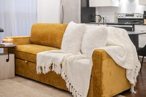 A mustard-colored sofa with a white throw blanket, a small table with a candle, and a kitchen in the background. - Stylish Retreat Near Crescent & Sainte-Catherine (Montreal)