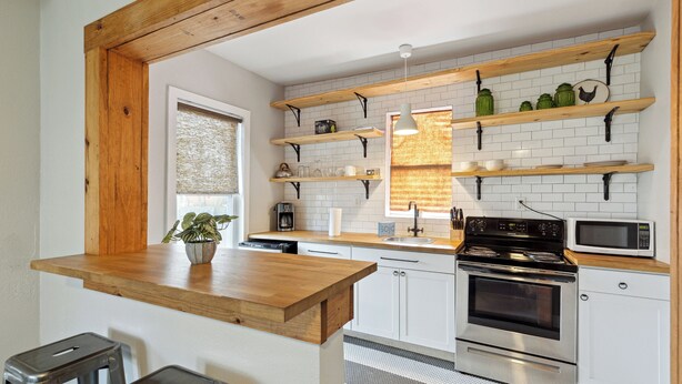 Una cocina moderna con armarios blancos, estantes de madera, encimera de madera y electrodomésticos de acero inoxidable, además de una planta en maceta.