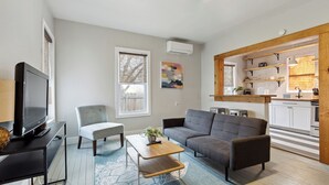 Una sala moderna con un televisor de pantalla plana, un sofá gris, un sillón blanco y una mesa de centro. Incluye una cocina con armarios blancos y estantes de madera.