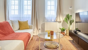 En moderne stue med sofa, sofabord i glas, fjernsyn og et stort vindue med gardiner.