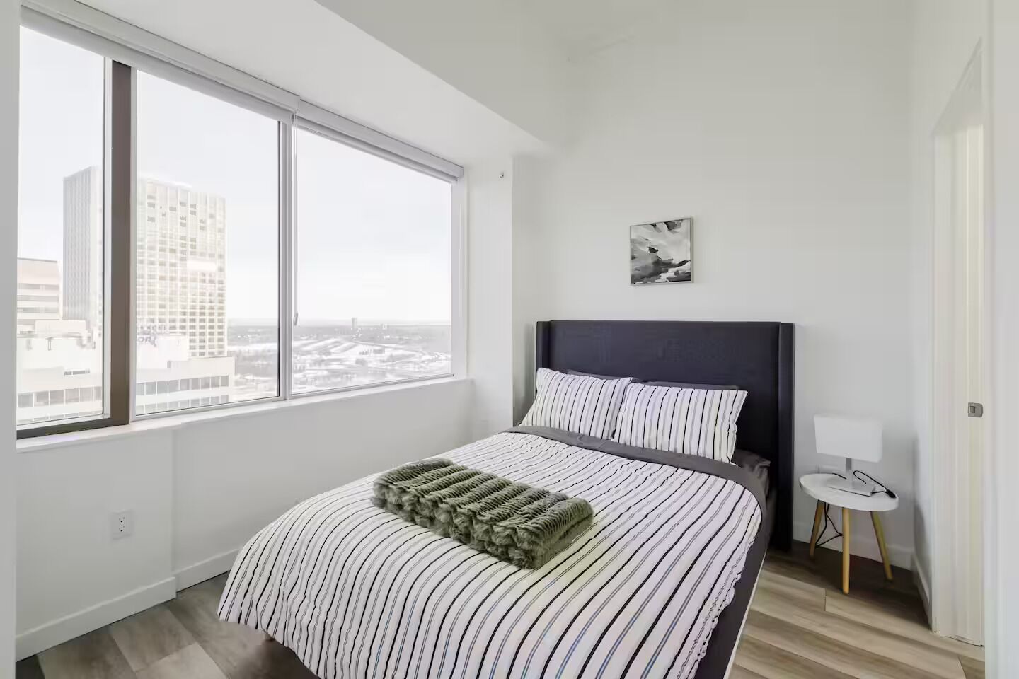 Une chambre moderne avec une grande fenêtre, un lit avec une literie à rayures, une tête de lit et une petite table de chevet avec une lampe.