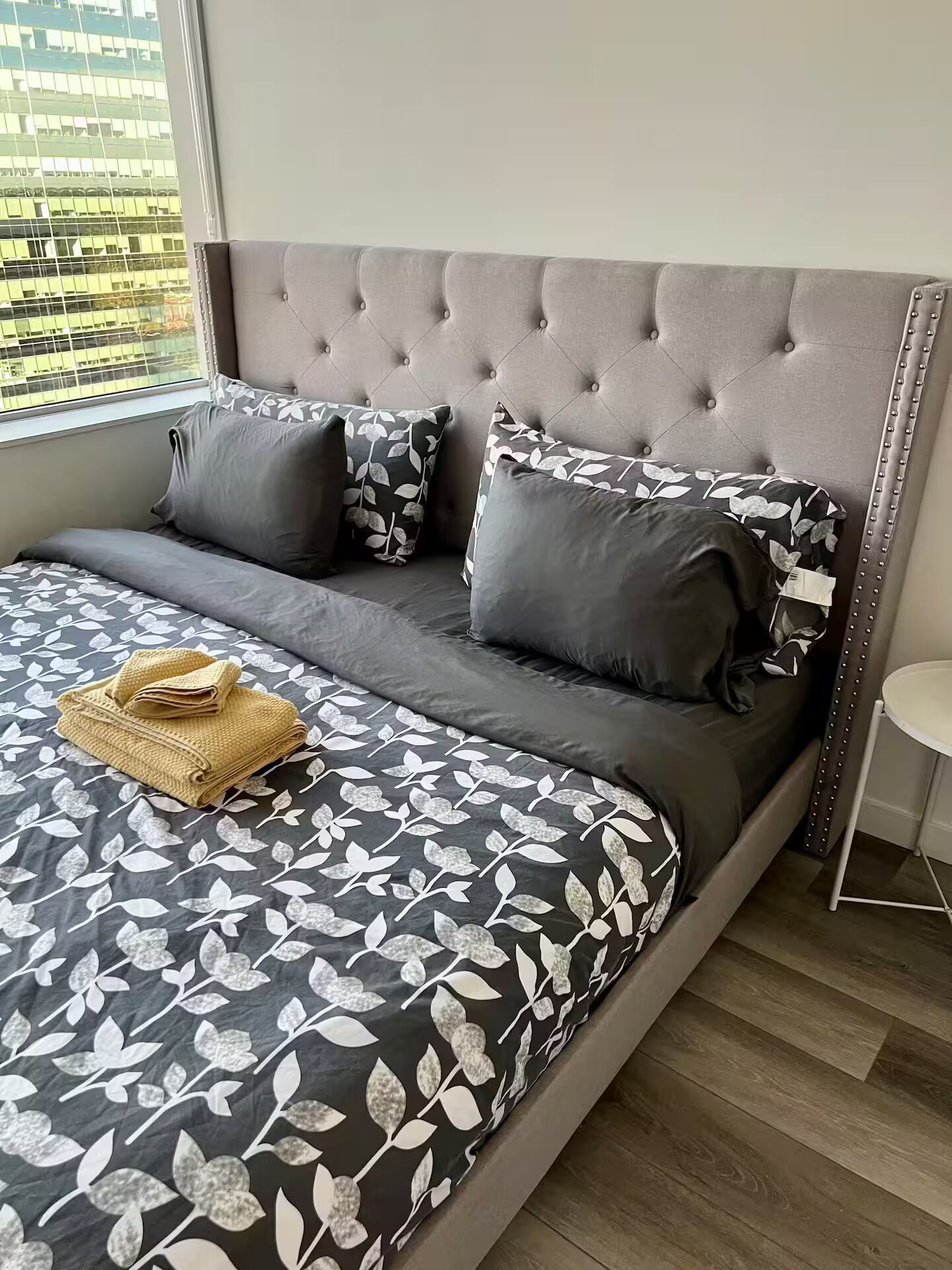 Een net opgemaakt bed met een grijs bekleed hoofdbord, beddengoed met een grijs-wit patroon, en een klein rond tafeltje ernaast.