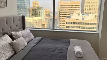 Une chambre moderne avec un grand lit, une vue sur le paysage urbain à travers de grandes fenêtres du sol au plafond, et une silhouette de la ville en arrière-plan.