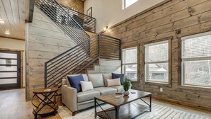 A spacious living room with a wooden staircase, a sofa set, a coffee table, and large windows. - Indoor Pool, Hot Tub! 'The Smoky Luxe Retreat' (Pigeon Forge)