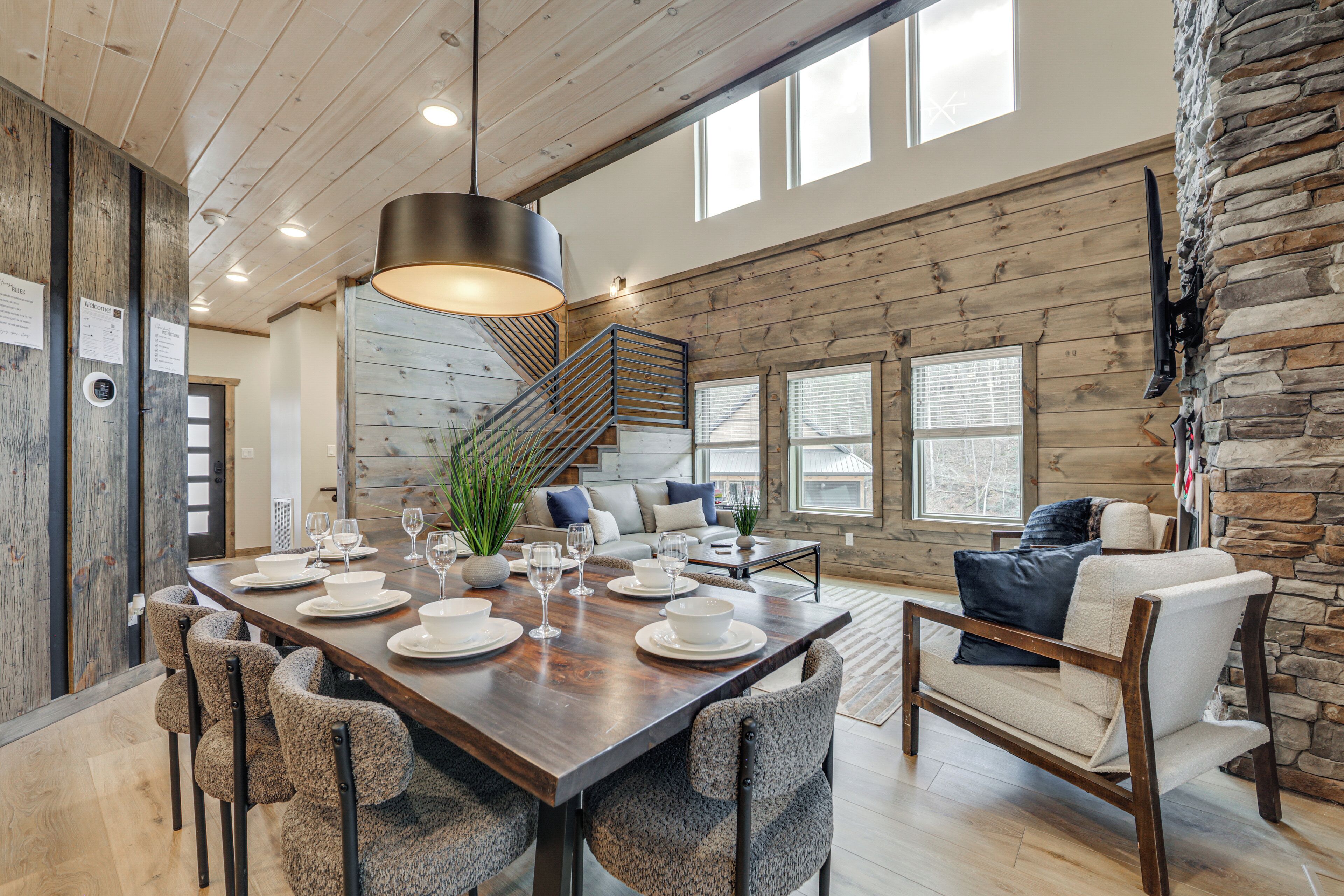 A spacious dining area with a long wooden table set for six, surrounded by upholstered chairs. A staircase with a metal railing leads to an upper level.