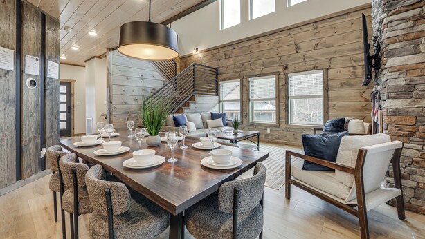 A spacious dining area with a long wooden table set for six, surrounded by upholstered chairs. A staircase with a metal railing leads to an upper level. - Indoor Pool, Hot Tub! 'The Smoky Luxe Retreat' (Pigeon Forge)
