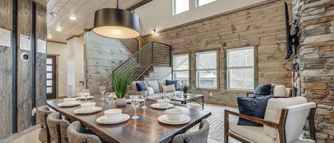 A spacious dining area with a long wooden table set for six, surrounded by upholstered chairs. A staircase with a metal railing leads to an upper level.