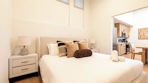 A bedroom with a large bed, bedside tables, a lamp, and a view into a kitchen area. - 2-bedroom apartment in terrific Montclair with AC and fitness room (Montclair)